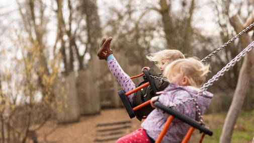Side-on view of two children with blonde hair and pink and brown winter coats are flying high on the swings in the Woodland Play Area.